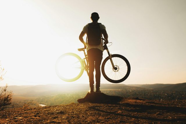 A cyclist stands silhouetted with a mountain bike on a hilltop during sunset, evoking adventure and freedom.
